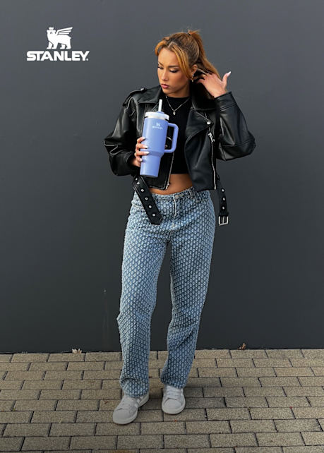 Female model wearing leather jacket and blue jeans while holding a blue Stanley cup
