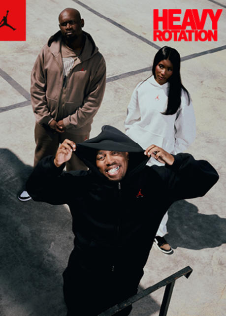 A promotional banner featuring three models standing on a grey concrete surface, viewed from a high angle. The model in the foreground adjusts a black hoodie, while the two models behind wear a brown track suit and a white hoodie. The top-left corner includes a red square with a black Jordan Jumpman logo, and the bottom-right corner features bold red text reading "HEAVY ROTATION."