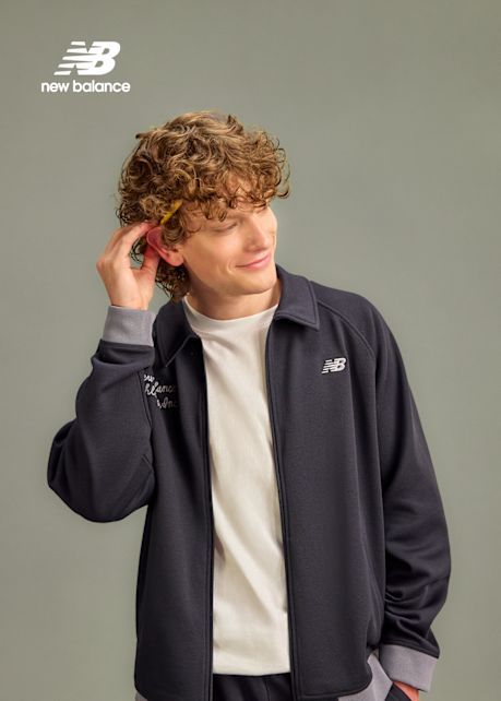 A medium-length portrait of a smiling person with curly light brown hair wearing a dark navy New Balance track jacket over a white t-shirt. Their right hand is reaching behind their ear. In the upper left corner, the New Balance logo is visible in white. The background is a flat, medium grey.