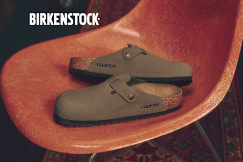 An overhead, slightly angled shot displays a pile of neutral-toned Birkenstock Boston clogs scattered across a detailed oriental rug. Prominently centered over the footwear is the bold, white "BIRKENSTOCK" brand logo.