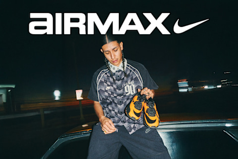 A split-screen Nike Air Max advertisement banner set outdoors under a clear sky. On the left side, two young men are looking toward the camera; one stands in the background wearing a black graphic t-shirt and camouflage pants while holding a flag pole, and the other sits in the foreground wearing a dark checkered short-sleeve button-up shirt featuring a small skull and crossbones logo. The word "AIRMAX" with the Nike swoosh is displayed in large white text across the top left. On the right side, a close-up, angled shot shows a black and mustard-yellow Nike Air Max sneaker with a visible air bubble in the heel and a small orange skull and crossbones graphic near the ankle.