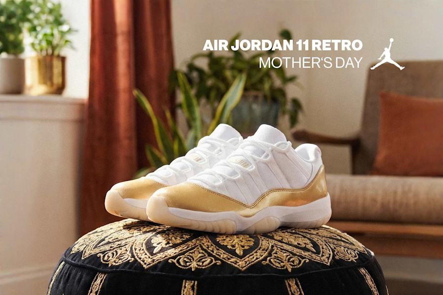 A promotional image for the "Air Jordan 11 Retro Mother's Day" sneakers. A pair of white low-top sneakers with a metallic gold patent leather mudguard are displayed on a black velvet ottoman with intricate gold embroidery. The background features a cozy living room setting with potted plants, a warm orange curtain, and a wooden armchair with a throw pillow. The Jumpman logo and the text "AIR JORDAN 11 RETRO MOTHER'S DAY" are overlaid in white at the top of the image.