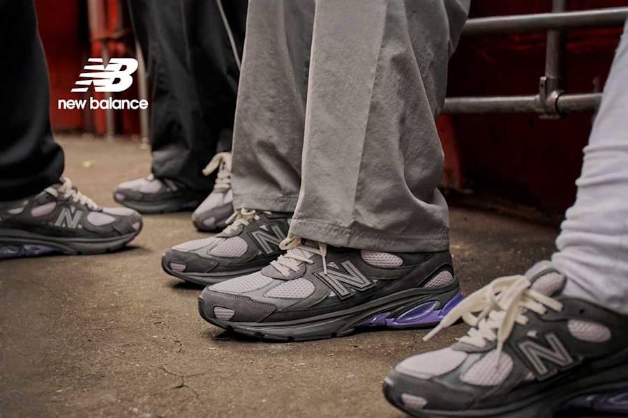 A man with a beard, tattoos, and a black baseball cap wears a grey New Balance tracksuit and grey 990v3 sneakers with purple accents while posing against a red wall and in a dark hallway; a supporting image shows a group of people wearing the same sneakers.