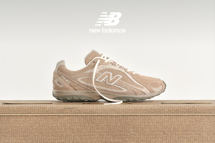 An eye-level, side-profile shot focuses on a person's lower legs and feet. They wear a tan New Balance sneaker with a large, white "N" logo on the side and cream-colored laces. Their white, ribbed crew socks feature a small, black New Balance logo. Above the socks, the bottom of a long, flared, brown and grey plaid skirt is visible. A white New Balance logo is in the top left corner. The background is a solid, light grey.