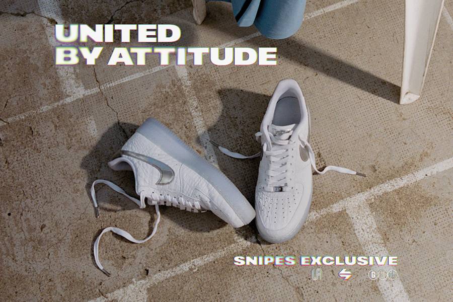 Top-down shot of a pair of white Nike Air Force 1 sneakers resting on a sunlit, textured concrete ground marked with white lines. The shoes feature a crinkled leather upper, metallic silver Swooshes, and translucent icy blue outsoles. One shoe lies on its side while the other stands upright. The top-left corner features the text "UNITED BY ATTITUDE" in a bold, outlined font, and the right side includes a vertical "SNIPES EXCLUSIVE" logo.