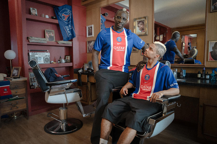 Two male models wearing PSG Homekit 26/27 Sakho jerseys and black pants, while being at the barber